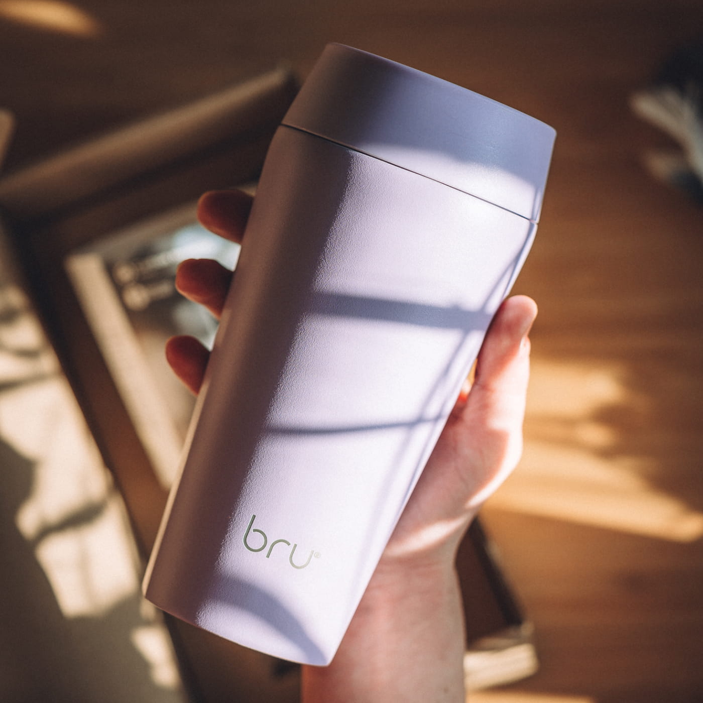 Shatterproof shell block: Hand holding a lilac bru travel mug in sunlight, showing the matt powder-coated 18/10 stainless steel outer shell
