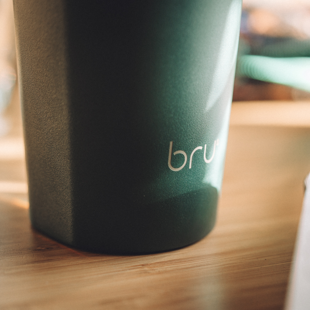 Side handles block: Close-up of a green bru travel mug on a wooden table showing the subtle built-in side handles for easy one-handed grip

